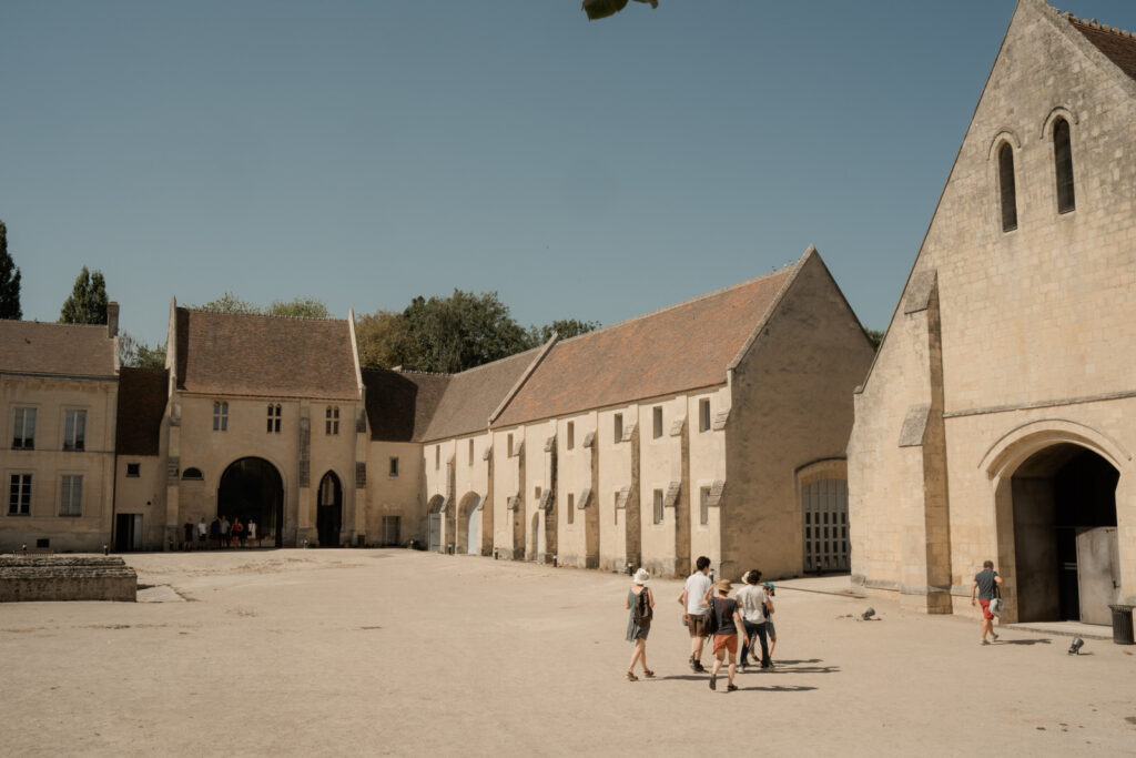 Vue d'ensemble de la cour pavée de l'abbaye d'Ardenne (IMEC) sous un ciel clair. L'architecture en pierre de Caen entoure l'espace. Plusieurs groupes de personnes déambulent et se dirigent vers les différentes entrées des bâtiments. À gauche, un grand calicot « AU PORTRAIT » est visible au-dessus d'une porte monumentale. Des buissons de lavande ornent le premier plan.